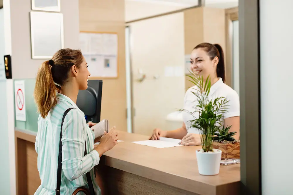 A smiling receptionist assisting a woman at the front desk of a medical or office setting, with a small potted plant on the counter.