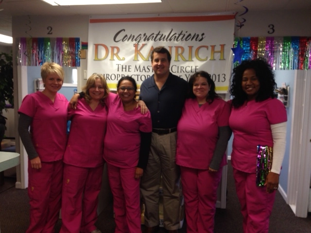 Chiropractor posing with clinic staff in pink scrubs in front of a congratulatory banner.