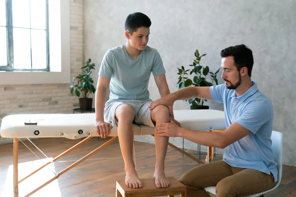 Physical therapist evaluates a young patient’s knee while the child sits on a treatment table during a physical therapy assessment in a bright, modern clinic.