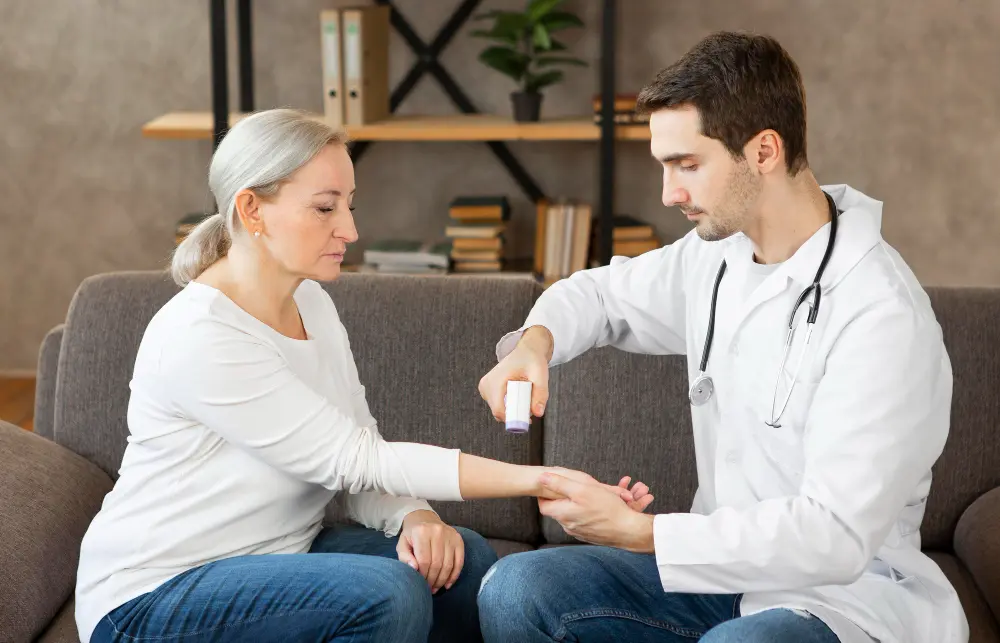 A healthcare professional in a white coat uses a handheld device to examine the wrist of an older woman sitting on a sofa in a home-like setting, with shelves and books in the background.