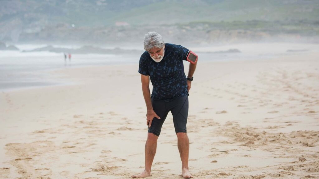 Athletic elderly man hunched over from sciatica pain running down his low back and hip on the beach