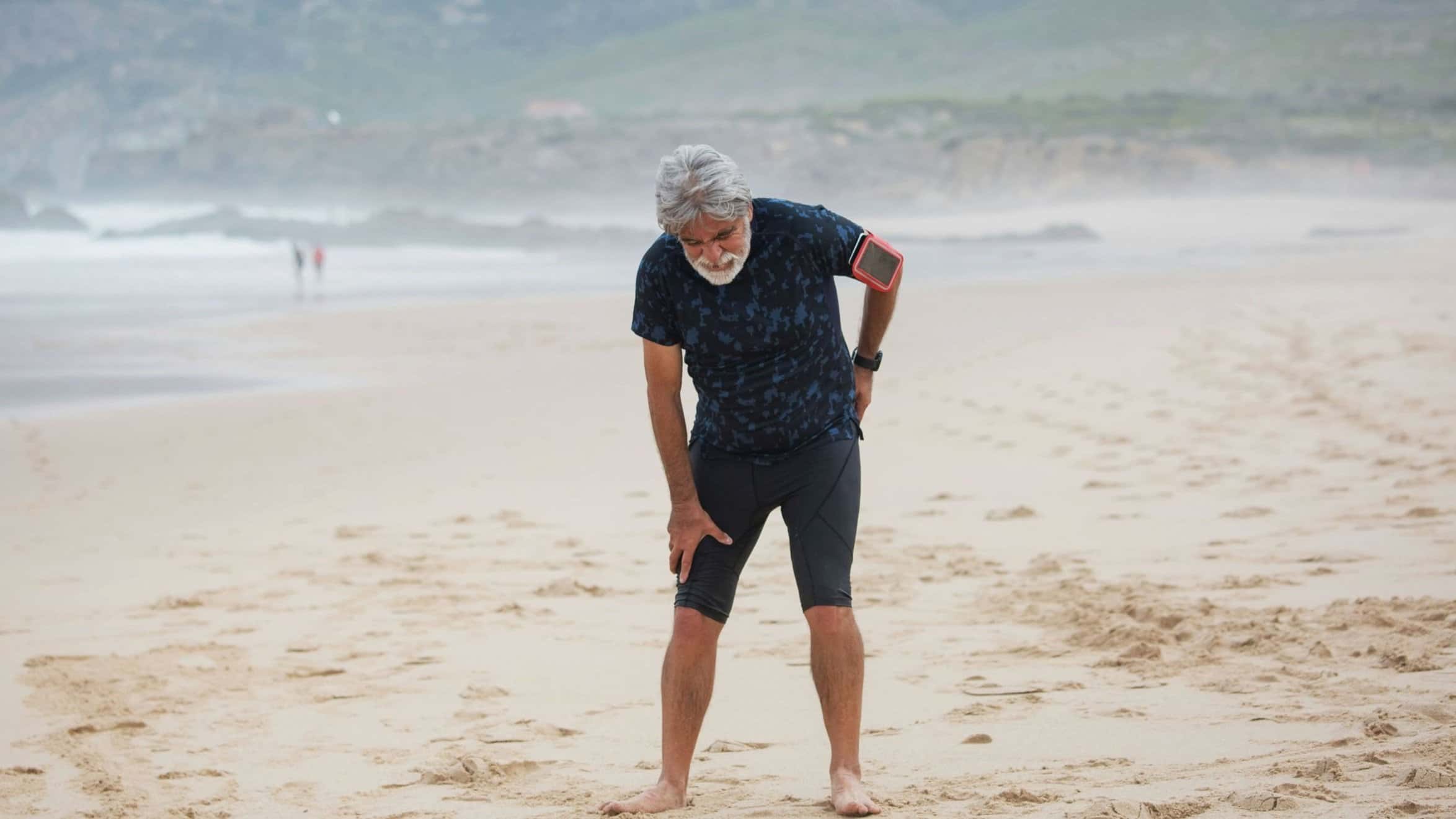 Athletic elderly man hunched over from sciatica pain running down his low back and hip on the beach